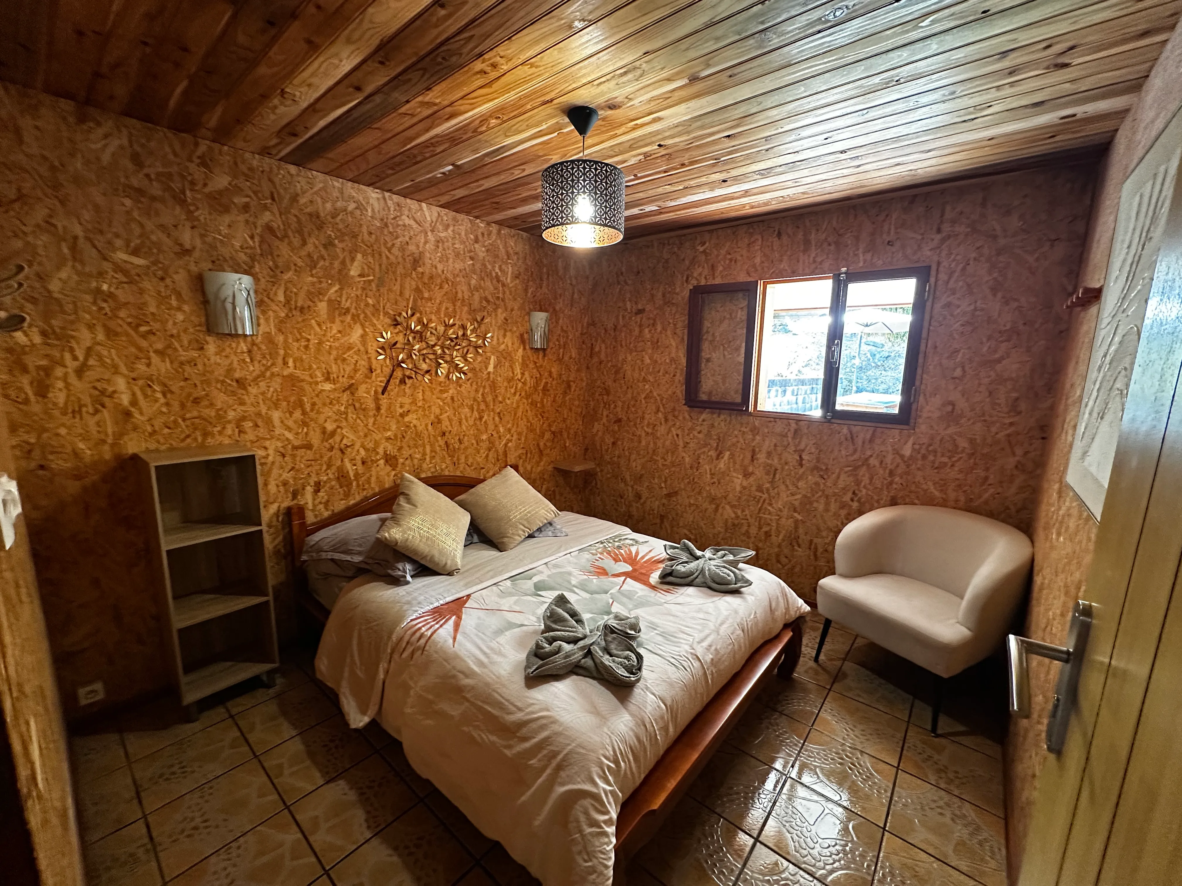 Chambre double n°1 du gîte : lit double avec coussins, murs et plafond en bois, fauteuil clair